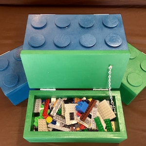 May include: A green wooden storage box shaped like a large building block, with a blue lid and a chain to hold the lid open. The box is filled with colorful toy building blocks. Additional blue and green blocks are visible.