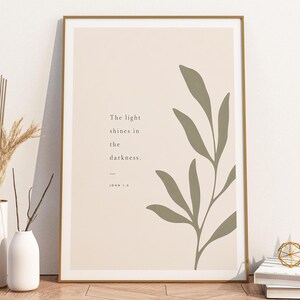 Könnte beinhalten: Ein rechteckiges gerahmtes Wandbild mit beigem Hintergrund, einer salbeigrünen botanischen Blattillustration und dem Text "The light shines in the darkness. — JOHN 1:5", der in einer dunkelgrauen Serifenschrift links zentriert ist.