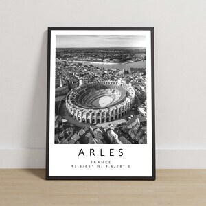 Op de afbeelding: Zwart-witte luchtfoto van het Romeinse amfitheater in Arles, Frankrijk. De tekst "ARLES FRANCE 43.6766° N, 4.6278° E" is onder de afbeelding gedrukt.