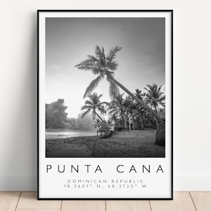Op de afbeelding: Zwart-wit ingelijste print met een strand met palmbomen. De print bevat de tekst "PUNTA CANA" en coördinaten. De afbeelding wordt weergegeven op een lichte houten vloer.
