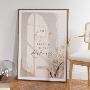 Peut inclure: Une affiche murale encadrée sur fond beige et crème avec le texte "THE light SHINES IN THE darkness" et "JOHN 1:5". Le design comprend une icône de soleil doré, un effet de bordure en papier déchiré et une illustration botanique de fleurs séchées.