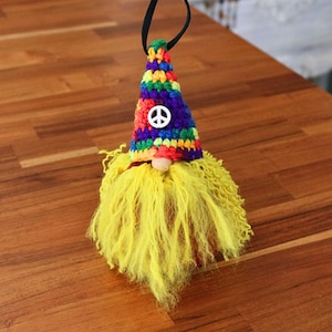 May include: A handmade gnome ornament with a colorful crocheted hat and a yellow yarn beard. The hat features a peace sign and a black hanging loop. The gnome's nose is a small wooden bead.