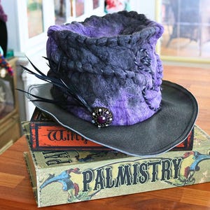 May include: A handcrafted, felted hat in shades of purple and gray, adorned with black feathers and a decorative jewel. The hat rests atop two vintage books with the titles "Witch" and "Palmistry" visible.