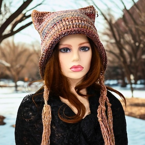 May include: A handmade crochet hat with cat ears in shades of brown, red, and blue. The hat has a brim and long, braided tassels. The hat is worn by a mannequin with long auburn hair and a black top. The background is a snowy park.