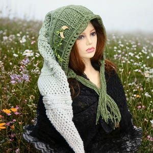 May include: A green and white crochet hood with a long, pointed extension. The hood features a wooden crescent moon and leaf accents. The background is a field of wildflowers.