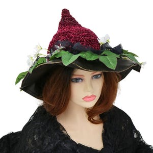 A crochet witch hat with a burgundy velvet pointed crown, black lace trim, and green leaves. The hat has a wide wired brim with green leaves and white flowers along the base of the crown. The stitching is intricately done and has a soft textural finish. A lush whimsical garden look for festivals, costumes, pagan events, or a festive Yule table centerpiece.