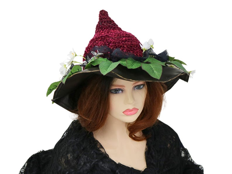 A crochet witch hat with a burgundy velvet pointed crown, black lace trim, and green leaves. The hat has a wide wired brim with green leaves and white flowers along the base of the crown. The stitching is intricately done and has a soft textural finish. A lush whimsical garden look for festivals, costumes, pagan events, or a festive Yule table centerpiece.