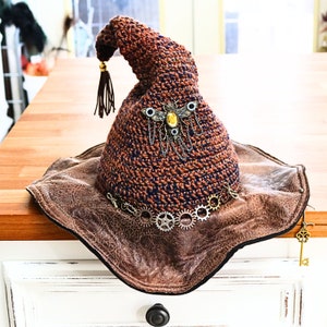 May include: A brown crocheted witch hat with a wide brim made of brown leather. The hat is decorated with a metal gear design and a jeweled insect.