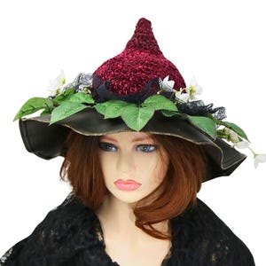 A crochet witch hat with a burgundy velvet pointed crown, black lace trim, and green leaves. The hat has a wide wired brim with green leaves and white flowers along the base of the crown. The stitching is intricately done and has a soft textural finish. A lush whimsical garden look for festivals, costumes, pagan events, or a festive Yule table centerpiece.