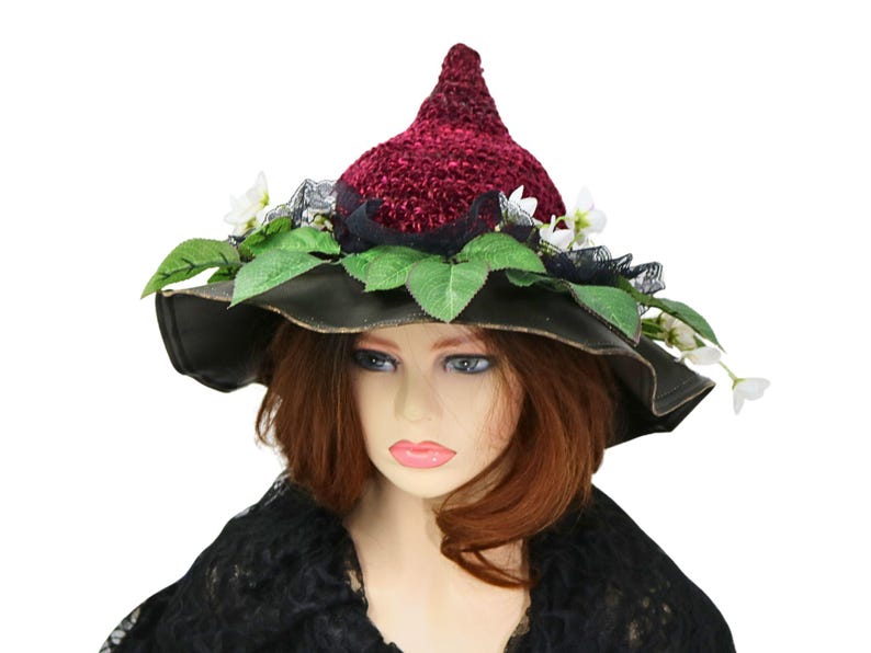 A crochet witch hat with a burgundy velvet pointed crown, black lace trim, and green leaves. The hat has a wide wired brim with green leaves and white flowers along the base of the crown. The stitching is intricately done and has a soft textural finish. A lush whimsical garden look for festivals, costumes, pagan events, or a festive Yule table centerpiece.