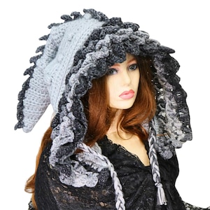 May include: A crochet hood featuring a light gray pointed crown with a textured spine and a dark charcoal gray ruffled border. The hood includes two braided ties in light gray and charcoal gray that hang down from the front edges of the piece.