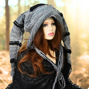 May include: A gray and black crocheted hood with two long, dangling side panels featuring horizontal black stripes. The hood is constructed from a textured yarn blend and includes long, braided yarn ties hanging from the front edges.