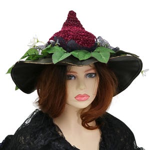 A crochet witch hat with a burgundy velvet pointed crown, black lace trim, and green leaves. The hat has a wide wired brim with green leaves and white flowers along the base of the crown. The stitching is intricately done and has a soft textural finish. A lush whimsical garden look for festivals, costumes, pagan events, or a festive Yule table centerpiece.