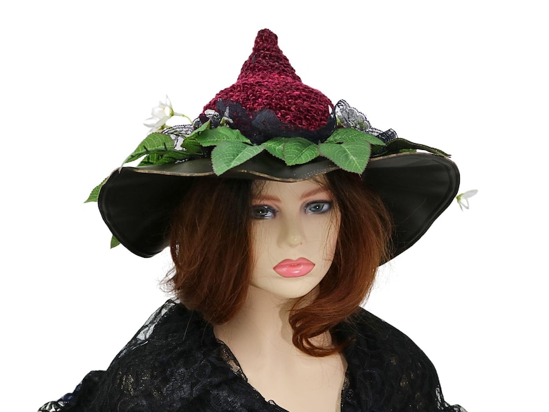 A crochet witch hat with a burgundy velvet pointed crown, black lace trim, and green leaves. The hat has a wide wired brim with green leaves and white flowers along the base of the crown. The stitching is intricately done and has a soft textural finish. A lush whimsical garden look for festivals, costumes, pagan events, or a festive Yule table centerpiece.