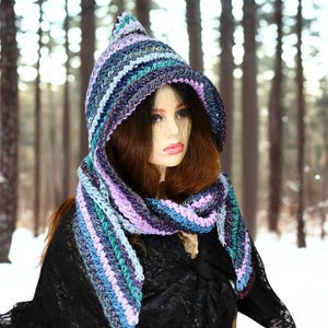 May include: A hooded scarf featuring a crochet knit pattern with horizontal stripes in shades of purple, lavender, teal, and dark blue. The accessory includes a pointed hood and long, draped ends that wrap around the neck, showcasing a textured yarn construction.