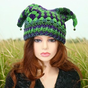May include: A green, purple, and blue crocheted hat with a pointed top and small dangling beads. The hat is worn by a mannequin with long auburn hair, against a backdrop of tall grass and a cloudy sky.