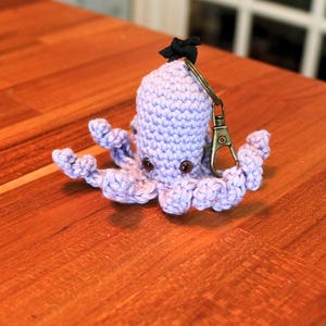 May include: A crocheted lavender octopus keychain with a black bow on top and a bronze-colored clasp. The octopus has eight curly tentacles and two brown button eyes. The keychain is sitting on a wooden surface.