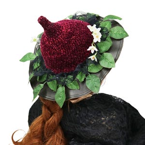 A crochet witch hat with a burgundy velvet pointed crown, black lace trim, and green leaves. The hat has a wide wired brim with green leaves and white flowers along the base of the crown. The stitching is intricately done and has a soft textural finish. A lush whimsical garden look for festivals, costumes, pagan events, or a festive Yule table centerpiece.