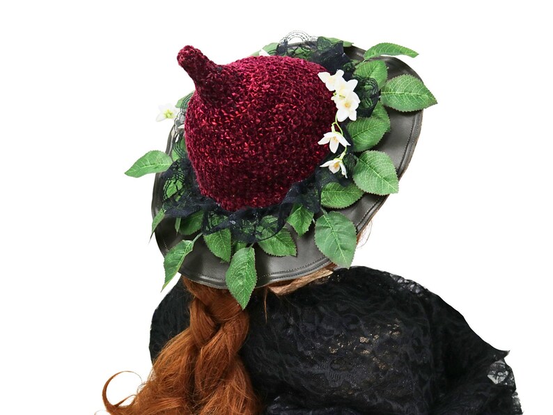 A crochet witch hat with a burgundy velvet pointed crown, black lace trim, and green leaves. The hat has a wide wired brim with green leaves and white flowers along the base of the crown. The stitching is intricately done and has a soft textural finish. A lush whimsical garden look for festivals, costumes, pagan events, or a festive Yule table centerpiece.