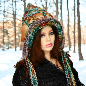 May include: A colorful, handmade crochet hat with a hood and long braids. The hat features a mix of green, brown, orange, and yellow yarn. The hat is worn by a mannequin. The background is a snowy forest.