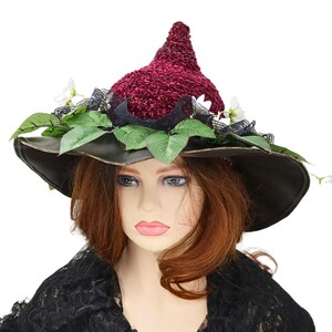 A crochet witch hat with a burgundy velvet pointed crown, black lace trim, and green leaves. The hat has a wide wired brim with green leaves and white flowers along the base of the crown. The stitching is intricately done and has a soft textural finish. A lush whimsical garden look for festivals, costumes, pagan events, or a festive Yule table centerpiece.