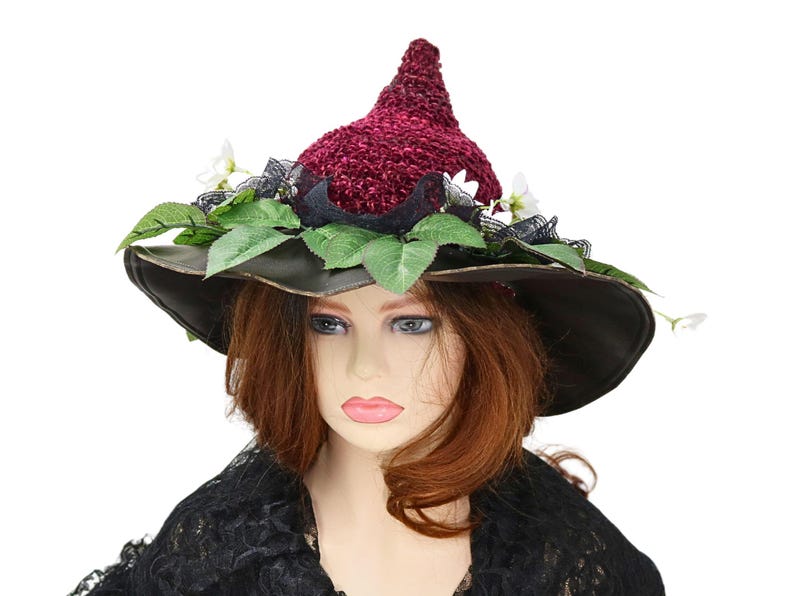 A crochet witch hat with a burgundy velvet pointed crown, black lace trim, and green leaves. The hat has a wide wired brim with green leaves and white flowers along the base of the crown. The stitching is intricately done and has a soft textural finish. A lush whimsical garden look for festivals, costumes, pagan events, or a festive Yule table centerpiece.