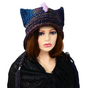 May include: A handmade, multi-colored crochet hat with pointed ear-like details. The hat features a mix of blue, purple, and brown yarns, with a small, light purple accent on top. Long, braided ties with white accents hang down the sides.