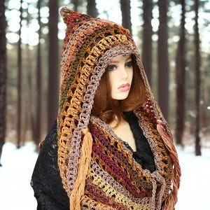 May include: A crochet hooded cowl featuring a variegated color palette of rust, brown, burgundy, and tan. The accessory has an open, textured stitch pattern with a pointed hood and long, dangling yarn tassels hanging from the front edges.