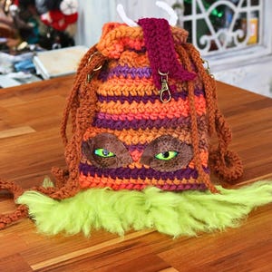 May include: A handmade crochet drawstring bag with a monster-like design. The bag features orange, purple, and brown stripes, with brown faux leather eyes and green irises. It has brown braided straps and a green fluffy bottom.