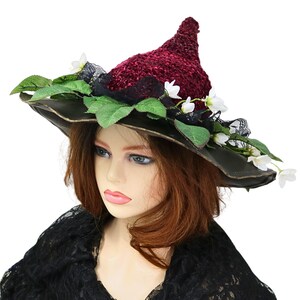 A crochet witch hat with a burgundy velvet pointed crown, black lace trim, and green leaves. The hat has a wide wired brim with green leaves and white flowers along the base of the crown. The stitching is intricately done and has a soft textural finish. A lush whimsical garden look for festivals, costumes, pagan events, or a festive Yule table centerpiece.