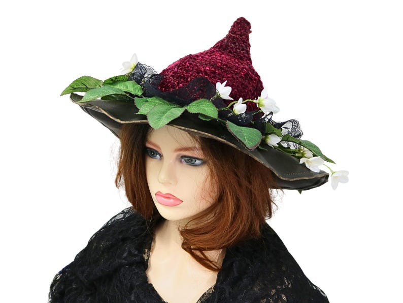 A crochet witch hat with a burgundy velvet pointed crown, black lace trim, and green leaves. The hat has a wide wired brim with green leaves and white flowers along the base of the crown. The stitching is intricately done and has a soft textural finish. A lush whimsical garden look for festivals, costumes, pagan events, or a festive Yule table centerpiece.