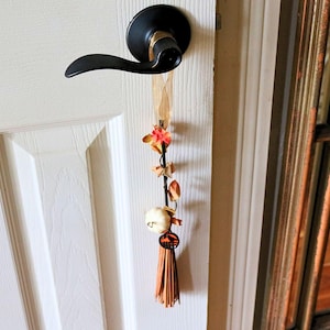 May include: A decorative door hanger with a black handle and a white door. The door hanger features a beige ribbon, orange and brown floral accents, a white ball, and a brown tassel. The door handle is black.