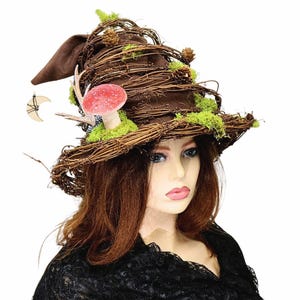 May include: A brown witch hat with a pointed crown, adorned with woven twigs, green moss, and a red mushroom. A small wooden crescent moon hangs from the side. The hat is a unique accessory for Halloween or costume events.