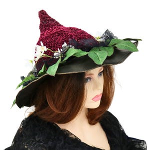A crochet witch hat with a burgundy velvet pointed crown, black lace trim, and green leaves. The hat has a wide wired brim with green leaves and white flowers along the base of the crown. The stitching is intricately done and has a soft textural finish. A lush whimsical garden look for festivals, costumes, pagan events, or a festive Yule table centerpiece.