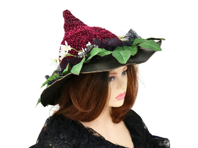A crochet witch hat with a burgundy velvet pointed crown, black lace trim, and green leaves. The hat has a wide wired brim with green leaves and white flowers along the base of the crown. The stitching is intricately done and has a soft textural finish. A lush whimsical garden look for festivals, costumes, pagan events, or a festive Yule table centerpiece.