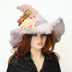 May include: A whimsical witch hat in shades of pink and gray, adorned with a cream-colored rose and small decorative pumpkins. The hat has a wide, ruffled brim and a pointed crown, perfect for Halloween or costume parties.
