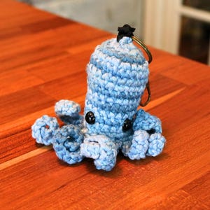 May include: A handmade blue and white crocheted octopus keychain with black button eyes and a black star-shaped accent. The octopus is sitting on a wooden surface.