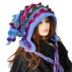 May include: A light blue crochet hood featuring a textured, scale-like pattern in shades of dark purple, forest green, and teal. The hood has a pointed, elongated shape and includes long, braided yarn ties in matching colors hanging from the front.