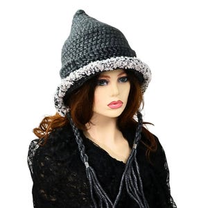 May include: A dark gray crocheted hat with a pointed top and a white, fluffy brim. The hat is part of a costume or fashion accessory, and is worn with a black shawl and gray braids.