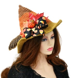 May include: A crochet witch hat in shades of brown, orange, and yellow, adorned with a black bow with orange accents, fall leaves, and a feather. The hat is decorated with a small pumpkin. The hat is on a mannequin.