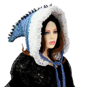 May include: A handmade blue and white crochet hood with a dragon scale design. The hood features a fluffy white trim around the face and a blue braided tie. The hood is designed to be worn as a costume or fashion accessory.