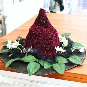 A crochet witch hat with a burgundy velvet pointed crown, black lace trim, and green leaves. The hat has a wide wired brim with green leaves and white flowers along the base of the crown. The stitching is intricately done and has a soft textural finish. A lush whimsical garden look for festivals, costumes, pagan events, or a festive Yule table centerpiece.