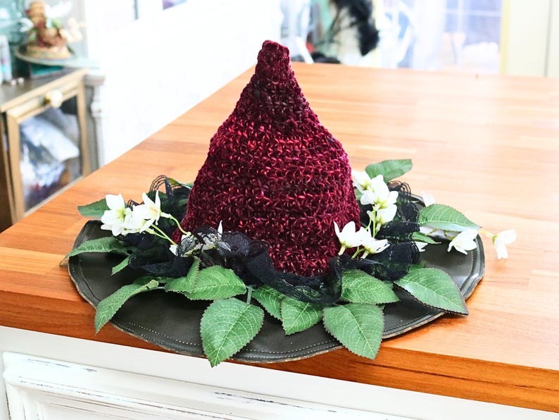 A crochet witch hat with a burgundy velvet pointed crown, black lace trim, and green leaves. The hat has a wide wired brim with green leaves and white flowers along the base of the crown. The stitching is intricately done and has a soft textural finish. A lush whimsical garden look for festivals, costumes, pagan events, or a festive Yule table centerpiece.
