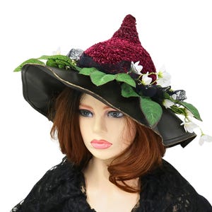 A crochet witch hat with a burgundy velvet pointed crown, black lace trim, and green leaves. The hat has a wide wired brim with green leaves and white flowers along the base of the crown. The stitching is intricately done and has a soft textural finish. A lush whimsical garden look for festivals, costumes, pagan events, or a festive Yule table centerpiece.