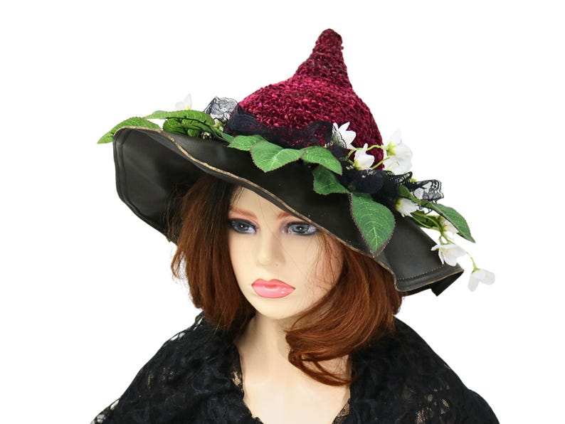 A crochet witch hat with a burgundy velvet pointed crown, black lace trim, and green leaves. The hat has a wide wired brim with green leaves and white flowers along the base of the crown. The stitching is intricately done and has a soft textural finish. A lush whimsical garden look for festivals, costumes, pagan events, or a festive Yule table centerpiece.
