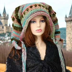 May include: A handmade, colorful crochet hat with a pointed hood and long, dangling tassels. The hat features a mix of pastel colors, including pink, blue, and green, with olive green accents. The hat is worn by a mannequin.