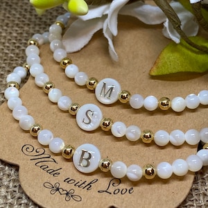 May include: Three white shell bead bracelets with gold accents. Each bracelet has a white charm with a gold letter, spelling out the word "B.S.M." The bracelets are on a brown card with the text "Made with Love".