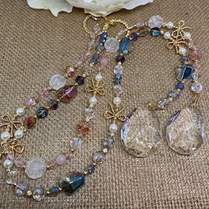 May include: A collection of beaded necklaces and keychains with iridescent, clear, and pearl-like beads. Gold-colored bow accents and teardrop-shaped crystal pendants are also featured. The jewelry is displayed on a textured, neutral-toned surface.