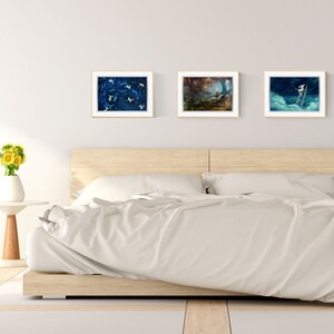May include: Three framed art prints hanging on a white wall above a wooden bed frame with white bedding. The prints feature abstract blue and white designs, a forest scene, and a figure climbing a ladder.
