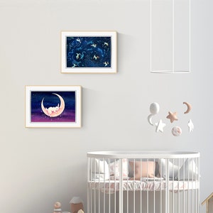 May include: Two framed illustrations of a white rabbit sleeping on a crescent moon and a constellation of stars in a dark blue sky. A white crib with a pink and white patterned bedding set is in the foreground.